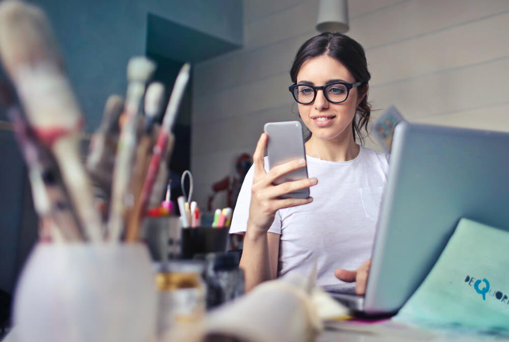 The Dream Client Quest - A Woman Looking at her phone with paintbushes in the foreground